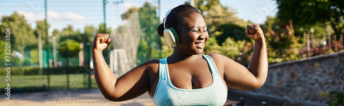 An African American woman in a sports bra top flexes her muscles confidently outdoors, showcasing body positivity and strength.