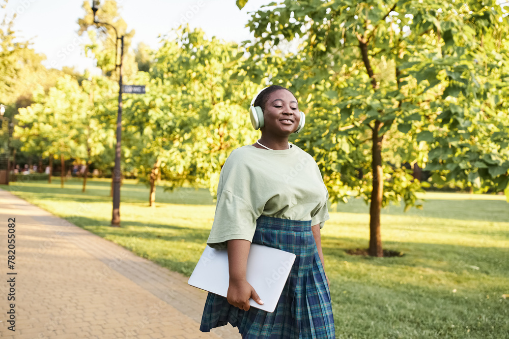 Fototapeta premium A plus size African American woman in casual attire walks confidently on a park sidewalk in summer.