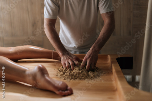 Cropped shot of unrecognizable massage practitioner male mixing heated sand on massage table for scrubbing treatment in luxury spa salon. Preparation to serenity of recreation in wellness center.