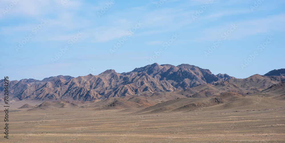 a wide desert with a vast, flat plain stretches out, covered with sparse, dry vegetation and rolling hills, leading up to a rugged mountain range with the light of the sun creating a play of shadows.