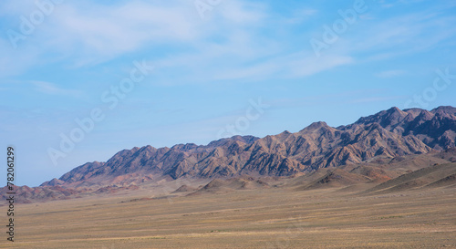 a wide desert with a vast  flat plain stretches out  covered with sparse  dry vegetation and rolling hills  leading up to a rugged mountain range with the light of the sun creating a play of shadows.