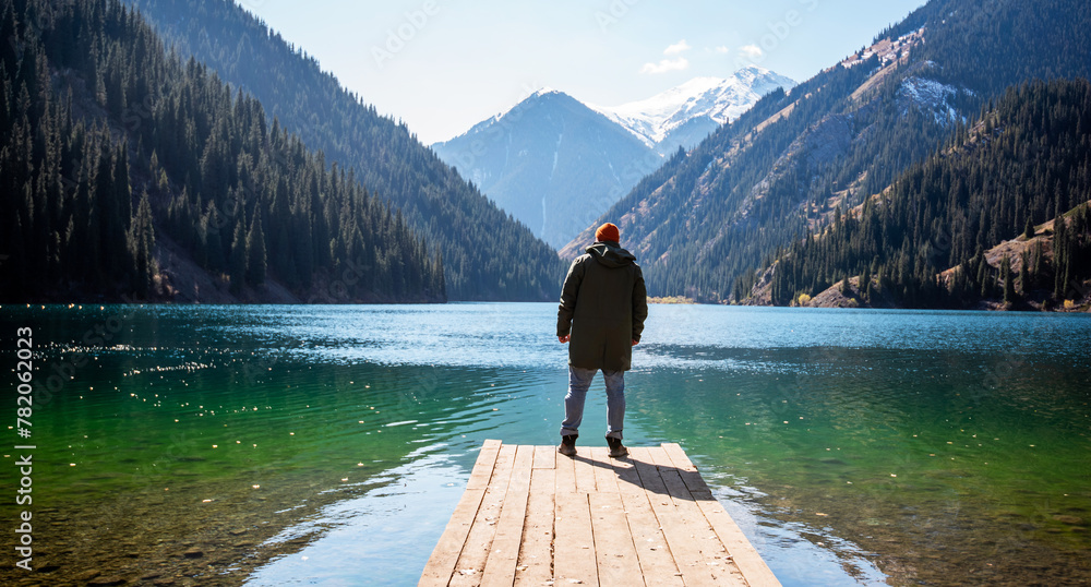 A man stands on a wooden pier looking out at a tranquil mountain lake, surrounded by forested slopes and snow-capped peaks under a clear blue sky. - obrazy, fototapety, plakaty 