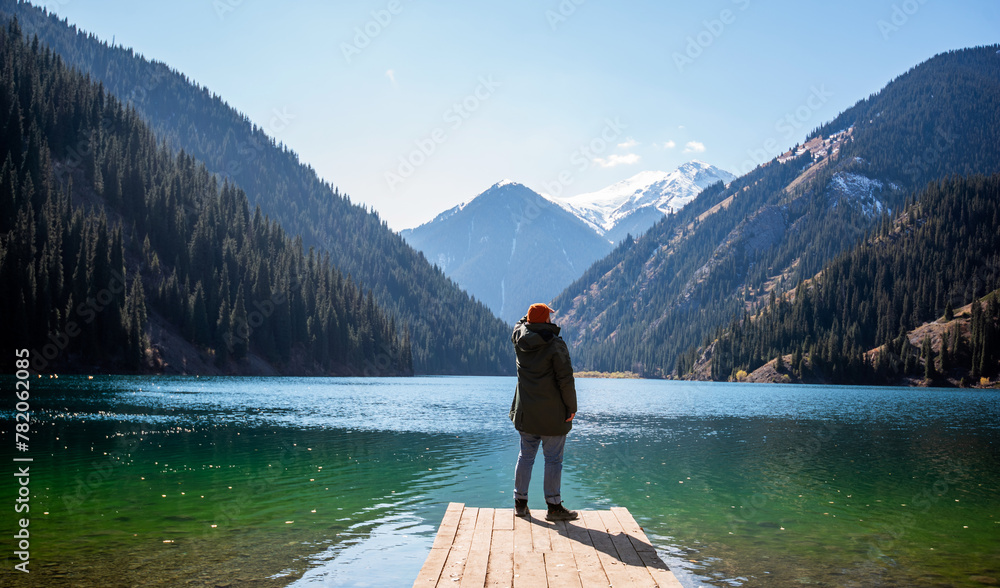 Obraz premium A man stands on a wooden pier looking out at a tranquil mountain lake, surrounded by forested slopes and snow-capped peaks under a clear blue sky.