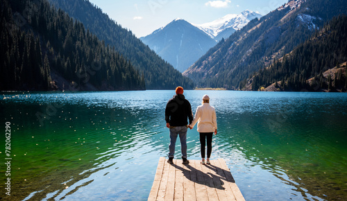 Obraz na plátně A man and woman hold hands on a dock, gazing at a mountain lake flanked by evergreen forests and snowy peaks beneath a clear sky