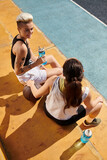 Two young female basketball players sitting on the ground, taking a break, enjoying each others company after a game.