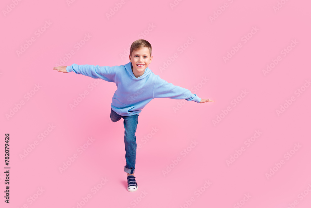 Full length photo of small boy stand one leg arms wings empty space isolated on pink color background Full length photo of small boy stand one leg arms wings empty space isolated on pink color background