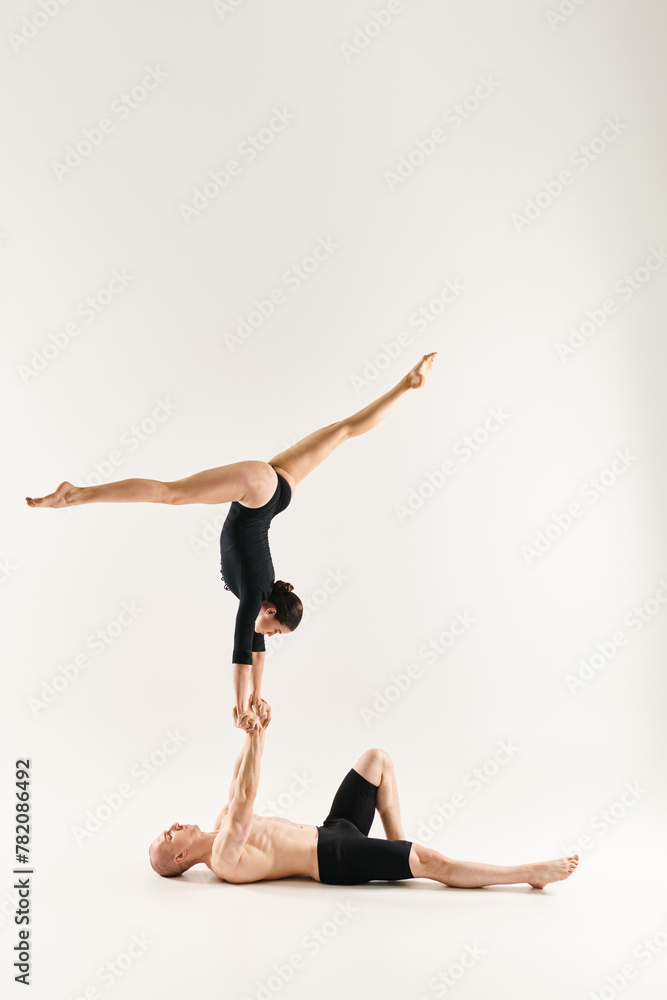A shirtless young man and a young woman gracefully performing a handstand together in a studio setting against a white background.