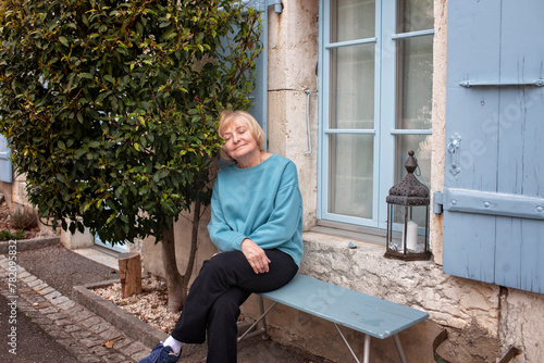 A contented elderly woman enjoys a peaceful moment on a bench outside her home, with a vibrant green tree and blue shutters in the background