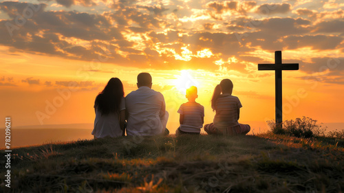 A religious family of four sits on a hillside looking at the sun as it sets with a cross next to them