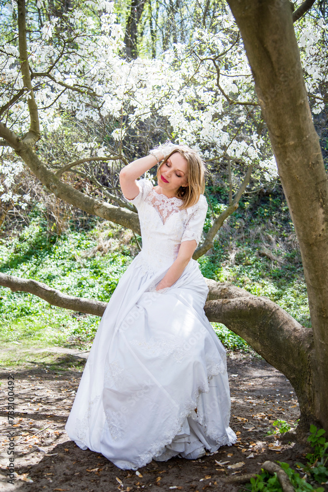 beautiful blonde smiling romantic bride in a white dress sitting on a branch in blossoming magnolia garden on sunny spring day