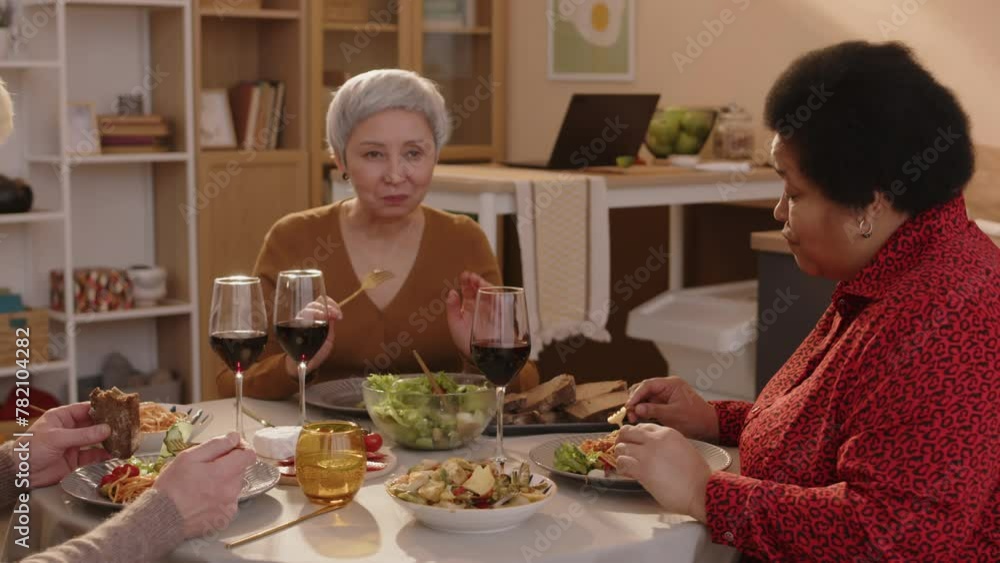 Medium shot of two multiethnic senior women and man chatting, drinking wine and eating dinner while getting together in cozy apartment Medium shot of two multiethnic senior women and man chatting, drinking wine and eating dinner while getting together in cozy apartment