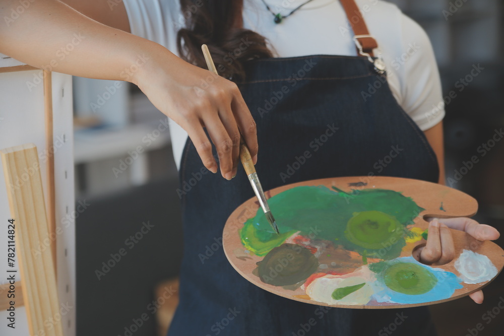 Obraz premium Cropped image of female artist standing in front of an easel and dipping brush into color palette