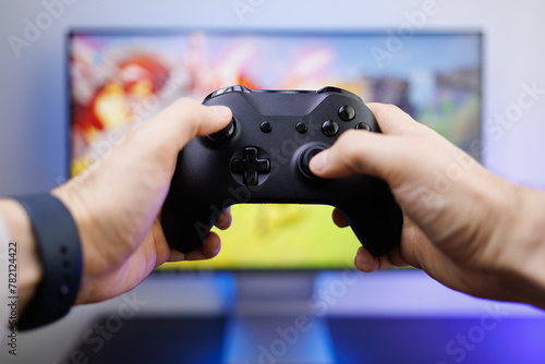 Hands hold a video game controller in front of the monitor. Video game gamepad in hand