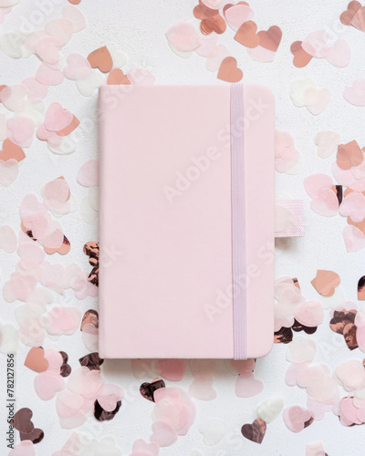 Pink hardcover notebook between pink hearts on white table top view, textbook mockup
