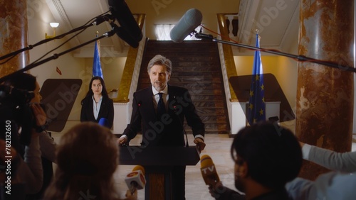Mature EU representative comes to press conference and gives interview for social media or TV news in government building hall. Confident consul political speech. Election coverage. Campaign speech.