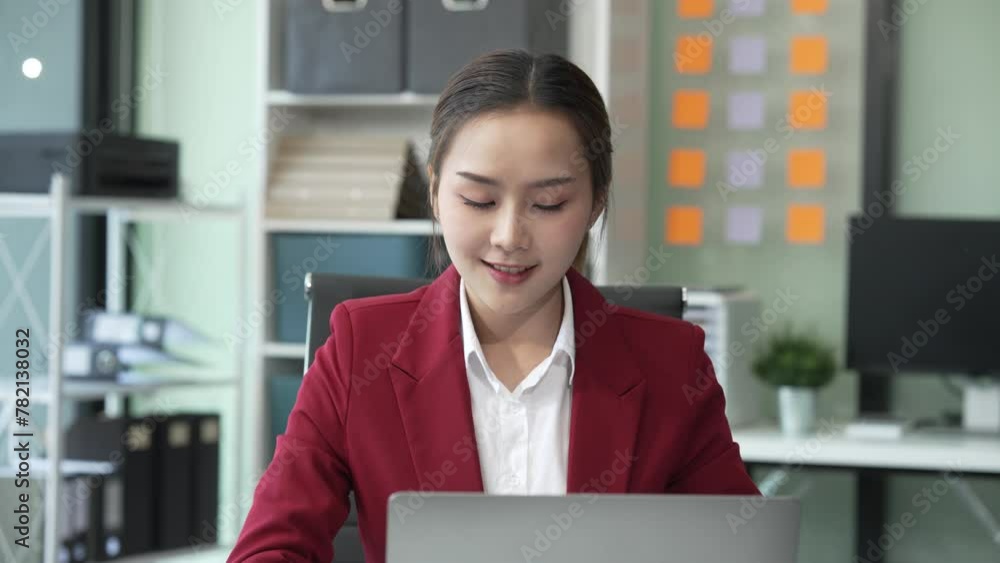 Asian young businesswoman, a Financial Advisor, works happily in her office. She uses her laptop and mobile phone to analyze charts and communicate financial strategies. Red formal suit Asian young businesswoman, a Financial Advisor, works happily in her office. She uses her laptop and mobile phone to analyze charts and communicate financial strategies. Red formal suit