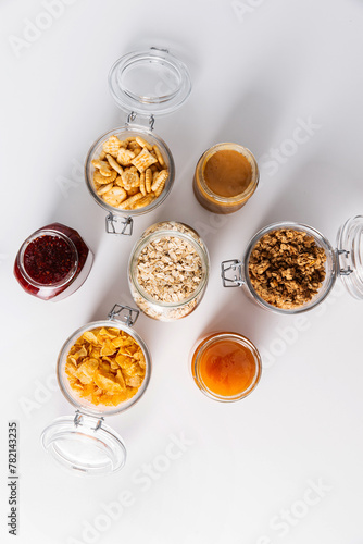 food storage and eating concept - close up of jars with oat, corn flakes, granola, cookies and spreads on white background