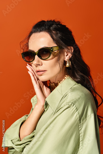 A fashionable woman in sunglasses and a green shirt poses against an orange background, exuding summertime vibes.
