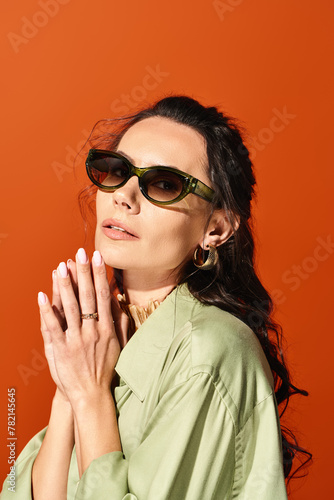 A fashionable woman wearing sunglasses and a green shirt poses confidently in a studio with an orange background. © LIGHTFIELD STUDIOS