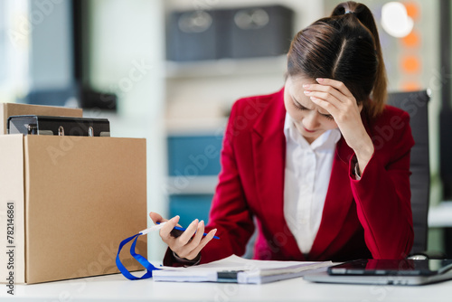 In the office, an Asian woman carefully signs her resignation letter, feeling stressed. She packs her belongings in cardboard boxes, preparing to leave the workplace. Asian people.