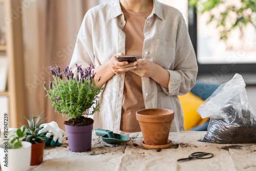 people, gardening and planting concept - close up of woman with smartphone and pot flower at home