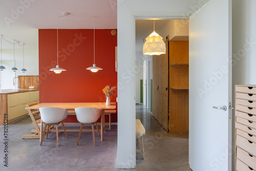Interior of a modern home in The Netherlands. Kitchen and dining area with design lamps and an orange wall. Hallway on the right.