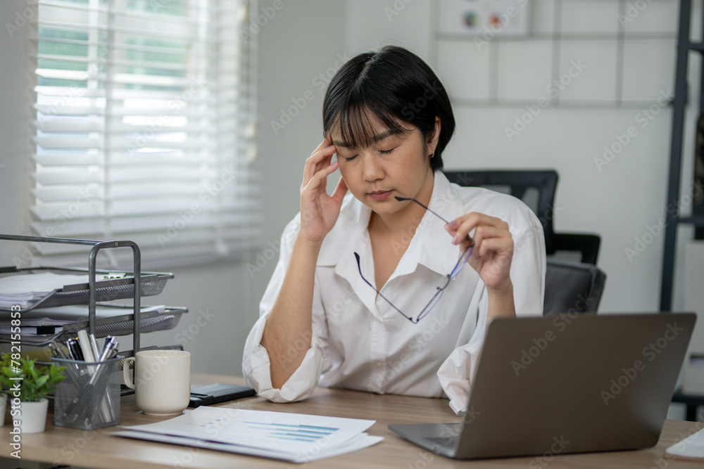 Exhausted female office worker feeling stressed, holding glasses, with laptop and documents on desk. Exhausted female office worker feeling stressed, holding glasses, with laptop and documents on desk.