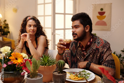 Young bearded Hispanic man with glass of tequila sitting by festive table next to his girlfriend and pronouncing toast in front of friends