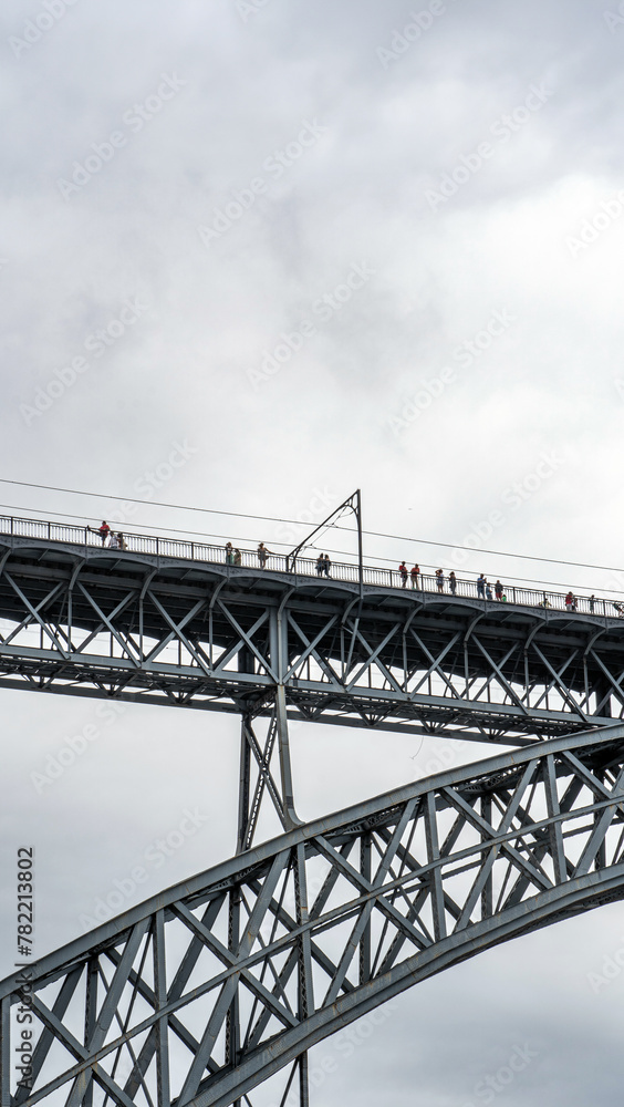 Tourists walking, taking photos and a woman posing from the upper platform of the Don Luis I steel bridge where the Porto metro passes with rain clouds.