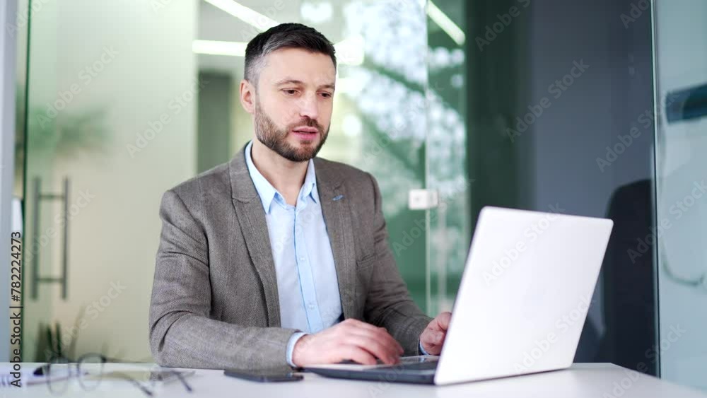 Confident businessman talking on a video call using a laptop computer sitting at a workplace in the office. A man worker has a business meeting, speaks remotely, communicates at an online conference Confident businessman talking on a video call using a laptop computer sitting at a workplace in the office. A man worker has a business meeting, speaks remotely, communicates at an online conference