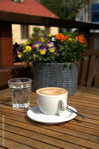 Cafe latte in a cup and glass of water on a table decorated with pansies in an ornamental flower pot 