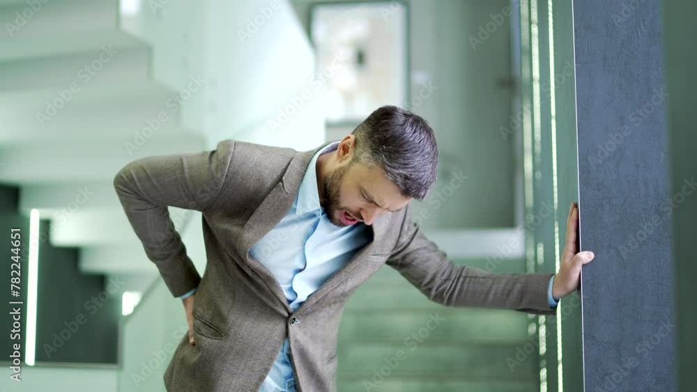 Tired businessman suffering from back pain while standing in corridor or hall of modern business center. Upset worker massages painful lower back muscles. Man in a jacket is exhausted and overworked Tired businessman suffering from back pain while standing in corridor or hall of modern business center. Upset worker massages painful lower back muscles. Man in a jacket is exhausted and overworked