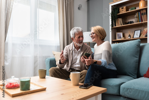 Senior couple, simple living. Older people man and woman full of love and respect, sitting at cozy home talking looking online funny videos on smartphone, enjoying their retirement and life. © Srdjan