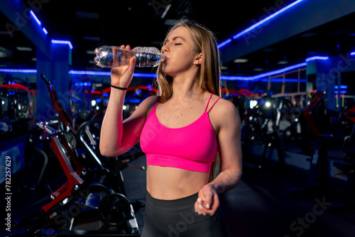 in the gym young beautiful girl with long hair in a pink top standing drinks water