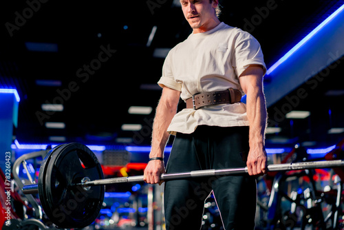 close up in the gym a young handsome guy in a T-shirt and a cap with a belt lifts a barbell with large athletic pancakes