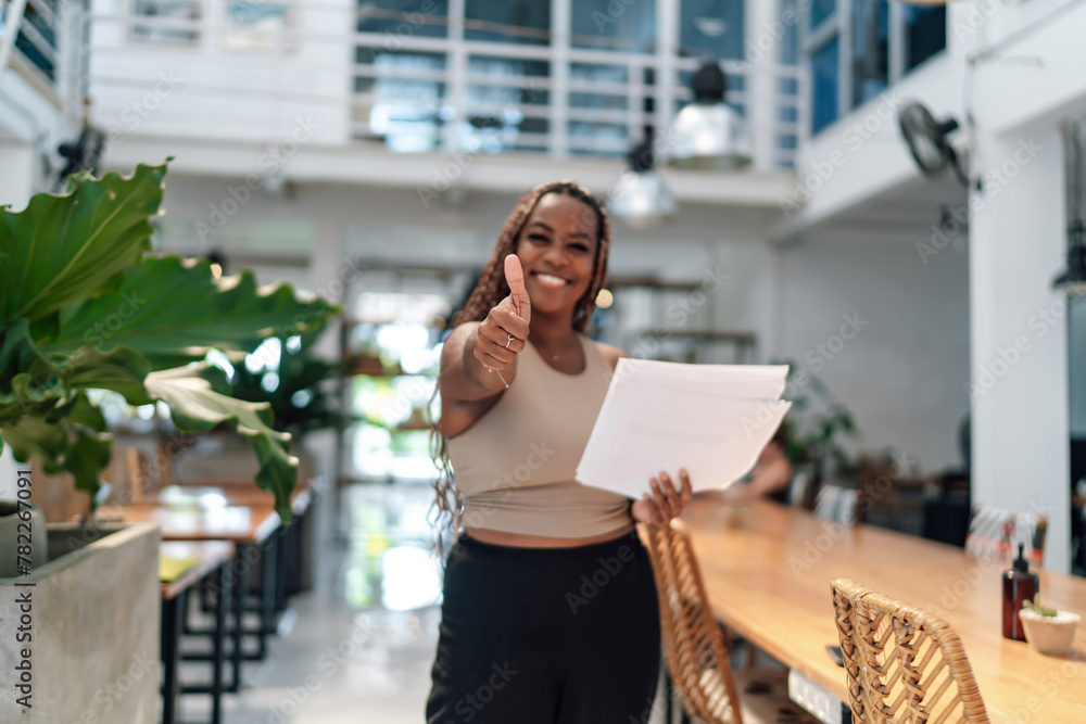 Fototapeta premium Young adult African-American female entrepreneur giving thumbs-up in a modern coworking space, holding documents, exuding confidence and professionalism.