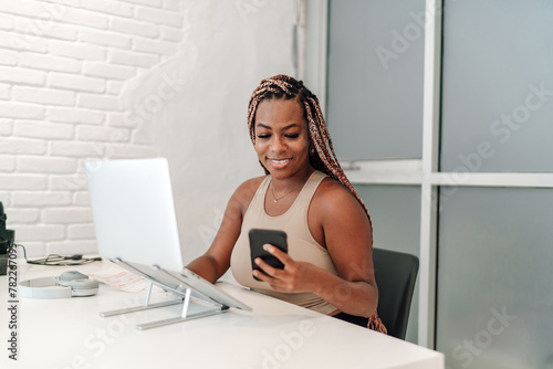 African-American woman entrepreneur in her 30s, business casual, working at white desk with a laptop computer while using smartphone application for managing tasks, cheerful in a well-lit office