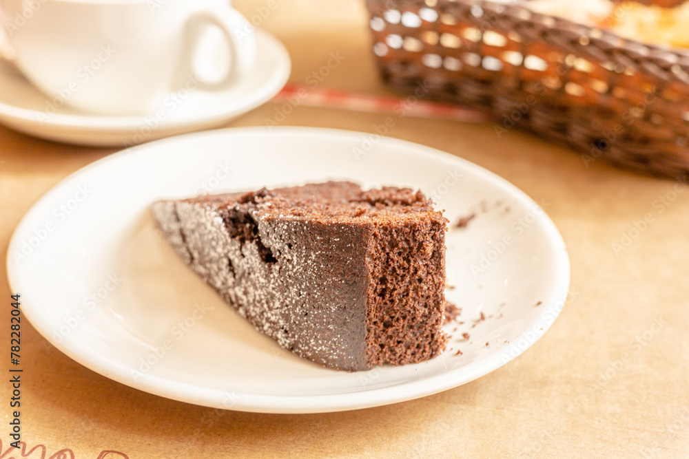 Piece of fresh homemade chocolate sponge cake on white plate, cup of tea or coffee on laid table