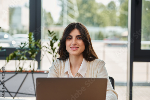 A businesswoman intensely focuses, sitting before a laptop in a modern office, embodying determination and commitment.