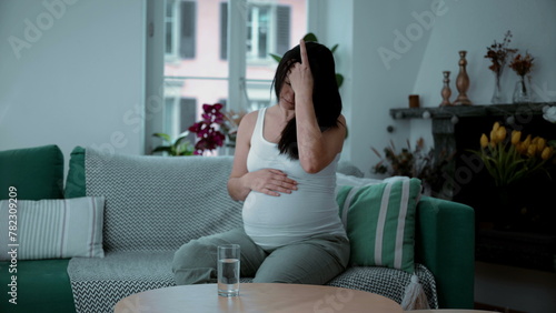 30s pregnant woman struggles with headache and pain seated at home sofa couch residence, maternal health concept