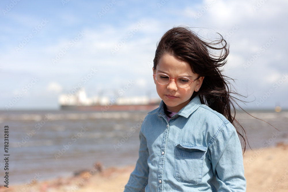 A little girl in glasses stands on the sea beach. Large ship for transporting goods in the background A little girl in glasses stands on the sea beach. Large ship for transporting goods in the background
