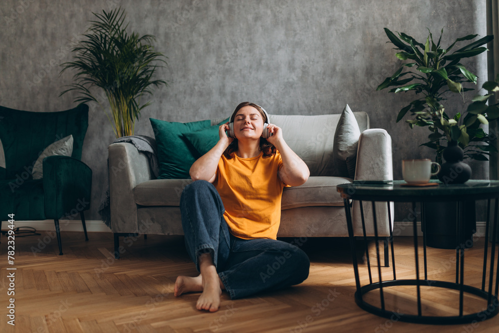 Smiling girl relaxing at home, she is playing music using a smartphone and wearing white headphones. Young woman hearing enjoying good audio sound lounge near couch at home, full body