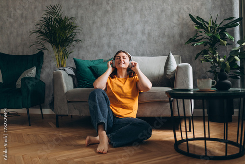 Obraz na płótnie Smiling girl relaxing at home, she is playing music using a smartphone and wearing white headphones. Young woman hearing enjoying good audio sound lounge near couch at home, full body w salonie