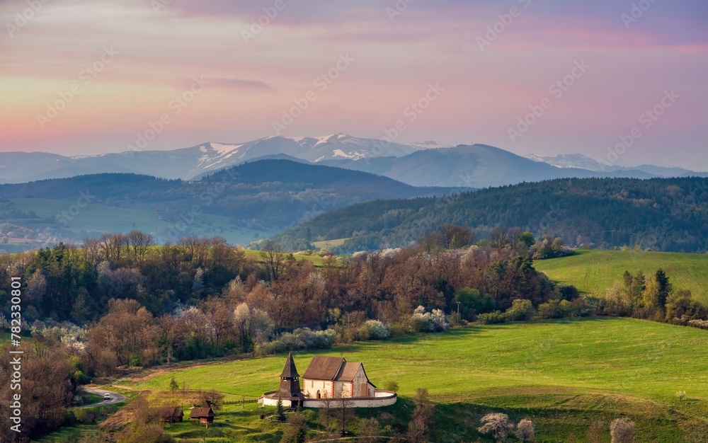 Picturesque spring mountain landscape with hills, meadows and romantic historic wooden building at colorful sunset. The village of Cerin near Banská Bystrica, Slovakia