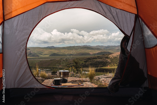 open orange tent with a view of camping pots and stove and black cup placed on the vegetation in the Andes mountain range with a lake background