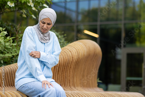 Obraz na plátně Muslim woman in hijab sitting outside, scratching her arm with a painful expression due to an apparent allergic reaction