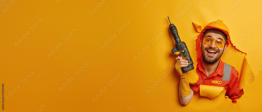 A delighted man in construction attire with a power drill pokes through a yellow wall, symbol for innovation A delighted man in construction attire with a power drill pokes through a yellow wall, symbol for innovation