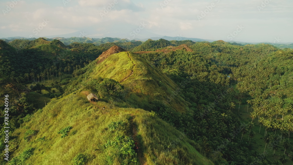 Green mount top aerial: travelers hiking path to building on hill top. Majestic Philippines nature landscape from mountain peak. Tourists climb at abudant greenery of tropical plants. Soft light shot