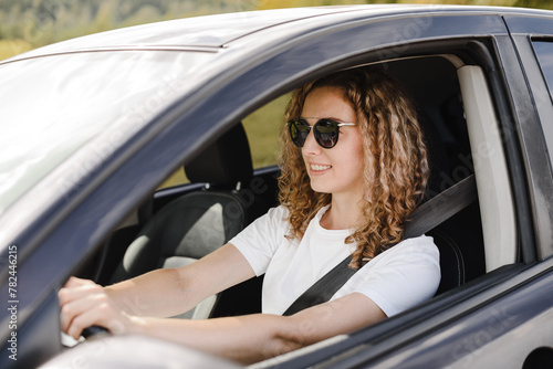 young beautiful smiling woman driving car, attractive caucasian woman in white t-shirt and black sunglasses © Anton Pentegov