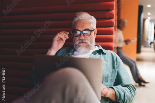 Thoughtful mature Caucasian man with a white beard using a laptop, seated on a red bench, with a senior woman in the background, in a modern office setting.